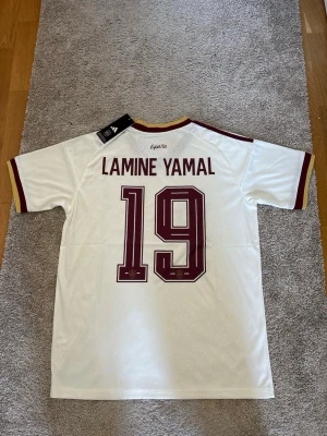 Vit Spanien fotbollströja Adidas - Säljer en vit Spanien fotbollströja från Adidas med Lamine Yamal och nummer 19 på ryggen. Tröjan har vinröda detaljer på ärmar och krage, samt Spaniens landslagsemblem och Adidas-logga på bröstet. Tillverkad i lätt och ventilerande material.