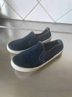 Mörkblå slip-on sneakers mesh - Säljer ett par mörkblå slip-on sneakers med mesh-överdel och vit sula. Skorna har elastiska paneler på sidorna för enkel på- och avtagning. Perfekt för dig som gillar en enkel och sportig stil. Materialet är lätt och andas bra.