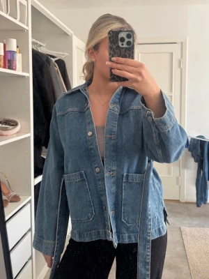 Blå jeansjacka med knytskärp - Säljer en blå jeansjacka med knytskärp i midjan och stora fickor framtill. Jackan har klassisk krage, knappar och raka ärmar. Perfekt för dig som gillar en avslappnad och trendig look.