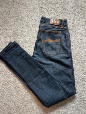 Nudie Jeans  - Snygga mörkblå jeans från Nudie Jeans med klassiska orange sömmar och ikonisk bakficka. Jeansen är i helt otroligt bra skick. Storleken är 29/34. Priset är ej hugget i sten🤗