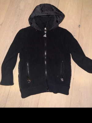 Svart cardiganjacka från Moncler - Svart stickad cardiganjacka från Moncler med dragkedja framtill och två fickor med zip. Jackan har en quiltad huva i nylon och Moncler-logga på ärmen. Perfekt för lager-på-lager och snygg till jeans.