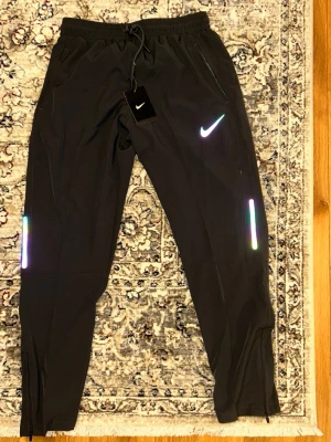 Gråa träningsbyxor från Nike - Snygga svarta träningsbyxor från Nike med reflexdetaljer. Helt nya. Storlek L men sitter mer som en S/M. Fraktas inom 48 timmar efter köp🚛