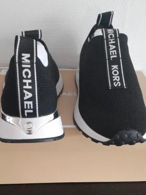 Svarta slip-on sneakers Michael Kors - Snygga svarta slip-on sneakers från Michael Kors med vit sula och silverdetalj på hälen. Skorna har tydliga logoband med texten 'MICHAEL KORS' på ovansidan och baktill. Ovandelen är i mesh-liknande syntetmaterial för en sportig look.