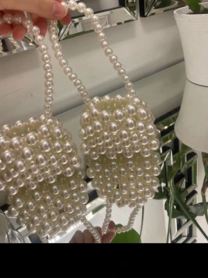 Handväska i pärlor med axelrem - Superfin handväska helt täckt av vita pärlor med matchande pärlhandtag. Väskan har en boxig form och är perfekt för dig som vill sticka ut med en unik accessoar. Passar till dig som gillar trendiga och lekfulla detaljer.