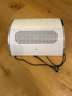 Nageldammsugare BeautyLushh vit - Nagelutsug från BeautyLushh i vit plast med svarta detaljer och silverfärgade kanter. Utrustad med ventilationsgaller och strömkabel, perfekt för att suga upp damm vid nagelbehandlingar.