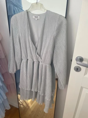Blågrå glitter omlottklänning från Kappahl - Blågrå omlottklänning från Kappahl med plisserad struktur och v-ringning. Klänningen har långa ärmar och volangdetaljer i nederkant😊