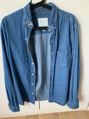 Blå jeansskjorta med knappar - Klassisk blå jeansskjorta i bomull med lång ärm, bröstficka och knäppning framtill. Skjortan har en avslappnad passform och tydliga sömmar för en tidlös denimlook. Perfekt att styla lager på lager eller bära som den är.