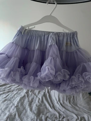 Ljuslila volangkjol från Dolly - Superfin ljuslila kortkjol från Dolly by Le Petit Tom med flera lager volanger i tyll. Kjolen har resår i midjan och ger en fluffig och lekfull look. Perfekt för dig som vill sticka ut med en söt och trendig stil.
