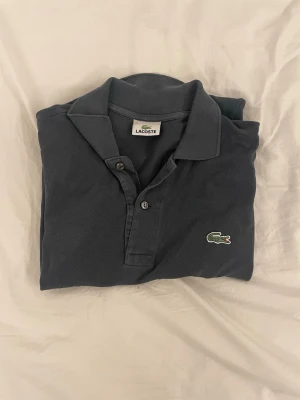 Mörkgrå Lacoste pikétröja - Säljer en mörkgrå pikétröja från Lacoste med klassisk krage och knappar framtill. Tröjan har det ikoniska krokodilmärket broderat på bröstet och är tillverkad i bomull. Perfekt för dig som gillar stilrena och tidlösa plagg.