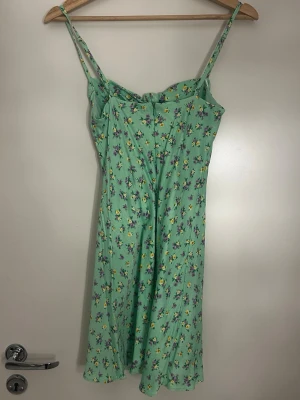 Grön blommig sommarklänning - Söt grön klänning med smala axelband och blommigt mönster i lila och gult. Klänningen har en feminin passform med lätt utställd kjol och knyt vid bysten. Perfekt för varma dagar och ger en fräsch look.