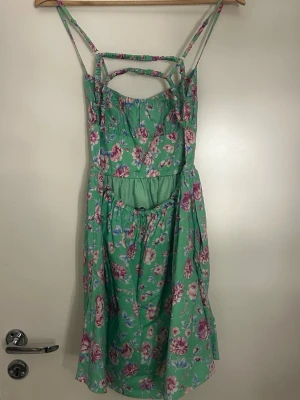 Grön blommig sommarklänning - Söt grön klänning med rosa och vita blommor, smala axelband och dekorativa banddetaljer vid bysten. Klänningen har en cut-out framtill och är i kort modell, perfekt för varma dagar. Tillverkad i ett lätt och luftigt bomullstyg.