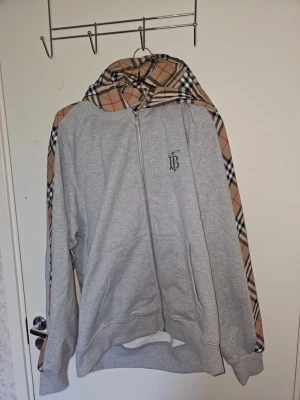 Grå Burberry hoodie - Snygg grå zip-hoodie från Burberry med klassiska beige, svarta och röda rutor på huva och ärmar. Dragkedja framtill, fickor och diskret TB-logga på bröstet. Perfekt för dig som vill ha en stilren men ändå unik hoodie med premiumkänsla. Den är oversized