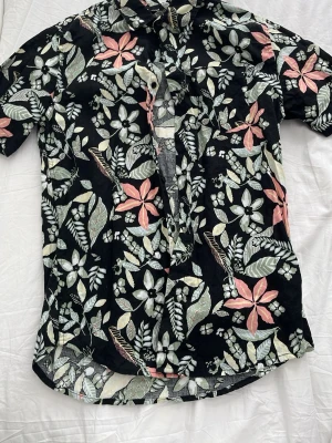 Blommig skjorta från 157, svart - Säljer en svart kortärmad skjorta från 157 med stort blommigt mönster i vitt, grönt och rosa. Skjortan har klassisk krage och knappar framtill. Perfekt för dig som vill sticka ut med en färgglad och somrig look. Tveka inte o höra av dig vid frågor😄