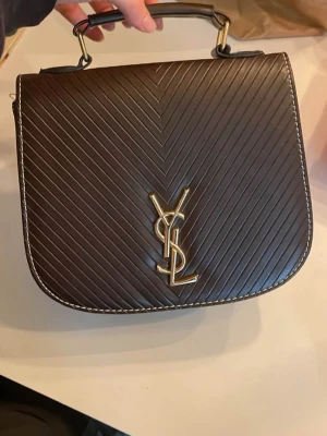 Brun handväska  - Snygg brun handväska guldfärgat logotyp på framsidan. Väskan har ett robust handtag och vita kontrastsömmar längs kanterna. Insidan är rymlig med ficka och guldfärgad kedja du kan ha över axeln. Perfekt accessoar för att lyfta din outfit.