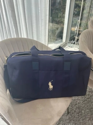 Mörkblå weekendbag från Polo Ralph Lauren - Snygg mörkblå weekendbag från Polo Ralph Lauren med klassisk gul broderad logga framtill. Väskan har både handtag och justerbar axelrem samt dragkedja i guldton. Perfekt för resor eller gymmet, tillverkad i slitstark canvas.