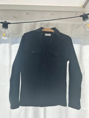 Svart overshirt från Jack and Jones  - Stilren svart overshirt från Jack and Jones  med klassisk krage och två stora bröstfickor med lock och knapp. Skjortan har raka ärmar och knäppning framtill. Perfekt att bära lager på lager för en avslappnad look.