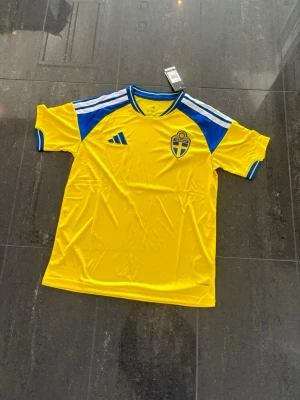 Svensk landslagströja Adidas gul/blå - Gul och blå fotbollströja från Adidas med Sveriges landslagsemblem på bröstet. Tröjan har vita ränder på axlarna, blå detaljer vid ärmar och hals samt klassisk Adidas-logga. Tillverkad i lätt och ventilerande material, perfekt för träning eller match.