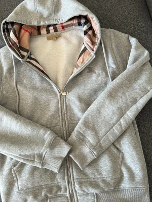 Grå hoodie från Burberry med dragkedja  - Snygg grå hoodie från Burberry med dragkedja och klassiskt rutigt foder i huvan. Mjuk bomullsblandning, ribbade muddar och fickor framtill. Perfekt för en stilren och avslappnad look.