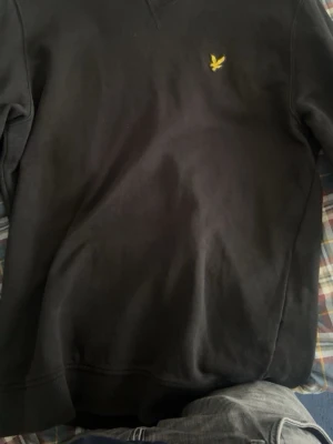 Svart sweatshirt från Lyle & Scott - En svart sweatshirt från Lyle & Scott med klassisk rund hals och det ikoniska gula fågel-logotypen broderad på bröstet. Tröjan har långa ärmar och ribbade muddar vid ärmslut och nederkant. Perfekt för en avslappnad och stilren look.