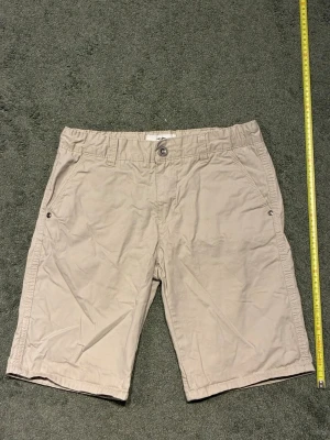 Beige shorts med fickor - Snygga beige shorts med normal passform. Shortsen har två sidofickor med knappdetaljer och två bakfickor med knappar. Klassisk midja och bälteshällor för enkel styling. Perfekta för varma dagar. Priset är ej huget i sten 