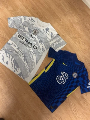 Manchester City och Chelsea matchtröjor - Säljer två fotbollströjor: en Manchester City bortatröja i vitt och ljusgrått kamouflagemönster från Puma med Etihad Airways-tryck i storlek S, samt en Chelsea hemmatröja i blått med gula detaljer och zigzag-mönster från Nike i storlek XS. Perfekta för match eller träning!