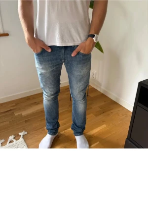 Blå slim fit jeans - Säljer ett par blå slim fit jeans med klassisk femficksdesign och snygga slitningar på låren. Jeansen har en modern passform som sitter tight längs benen och är tillverkade i denim. Perfekta för dig som gillar en stilren och avslappnad look.
