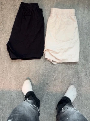 Svarta och beige shorts med snörning - Två par shorts i svart och beige med elastisk midja och snörning. Båda har bakficka och är tillverkade i ett mjukt bomullsmaterial. Perfekta för varma dagar och enkel stil.