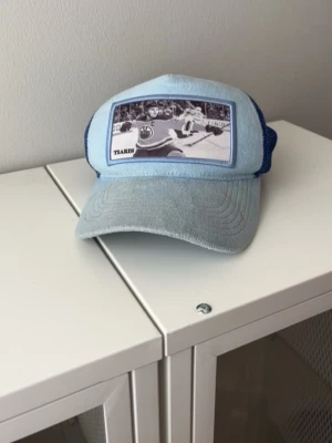 Blå keps med hockeymotiv från Tiaris - Cool ljusblå keps från Tiaris med mesh på sidorna och ett stort patch med hockeymotiv framtill. Perfekt för dig som gillar sport och vill ha en unik accessoar. Materialet är en blandning av tyg och mesh, vilket gör kepsen både luftig och bekväm.