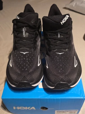 Svarta Hoka Clifton 9 sneakers - Snygga svarta Hoka Clifton 9 sneakers med vit logga och detaljer. Ovandelen är i mesh för bra ventilation och skorna har en chunky sula för extra komfort. Perfekta för dig som gillar streetstyle och sportig look.