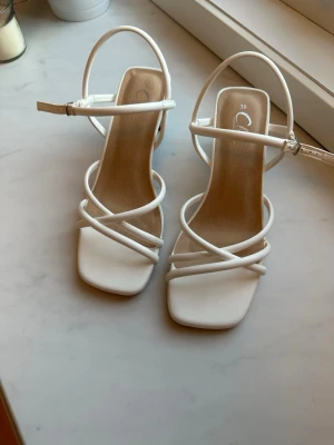 Vita sandaler  - Säljer ett par vita sandaler med trendiga smala remmar och fyrkantig tå. Sandalerna har en justerbar ankelrem och är tillverkade i syntetmaterial med en ljus innersula. Perfekt för dig som gillar stilrena och moderna skor. Använda kanske 5 gånger, några slitningar finns (syns på bild)