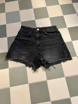 Svarta högmidjade jeansshorts - Säljer ett par svarta högmidjade jeansshorts med råa, fransiga kanter och klassisk femficksdesign. Shortsen har knapp och dragkedja framtill och är perfekta för en avslappnad och trendig look.
