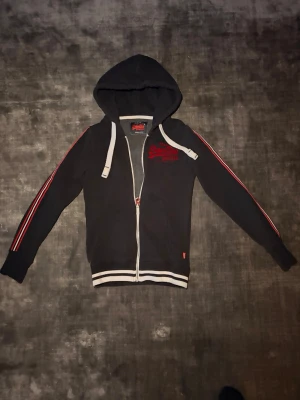 Superdry zip hoodie - Mörkblå hoodie från Superdry med dragkedja framtill. Snygga röda ränder längs ärmarna och röd broderad logga på bröstet. Vita snören i luvan och randig mudd nedtill som ger en sportig look. Perfekt till vardag eller lager-på-lager under kallare dagar.  • Märke: Superdry • Färg: Mörkblå med röda detaljer • Modell: Zip-hoodie med luva • Skick: mycket bra (se bilder) • Detaljer: Dragkedja, fickor, broderad logga  Säljs eftersom den inte kommer till användning längre. Hör gärna av dig🙌