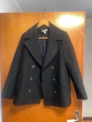 Svart skepparkavaj från H&M - Klassisk svart skepparkavaj från H&M med dubbelknäppning och breda slag. Jackan har två snedställda fickor och är helfodrad med svart tyg. Stilig och tidlös modell som passar perfekt till höst och vinter.