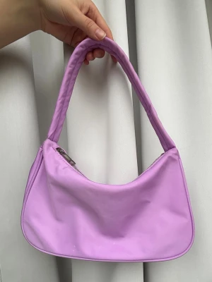 Ljuslila handväska i syntet - Trendig ljuslila handväska med mjuk, rundad form. Väskan har dragkedja upptill och är tillverkad i ett slätt syntetmaterial. Perfekt för dig som vill addera en färgklick till din outfit.