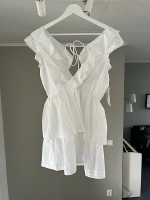 Vit omlottklänning med volang - Söt vit kortklänning med omlott och volangdetaljer. Klänningen har snörning i ryggen och är ärmlös med breda axelband. Tillverkad i ett lätt bomullstyg, perfekt för varma dagar. Romantisk och luftig design som ger en fräsch sommarkänsla.