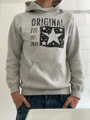 Scndi hoodie - Snygg grå hoodie med svartvit stjärntryck och texten 'ORIGINAL FIT EST. 2024' på bröstet. Hoodien har en klassisk känguruficka framtill och justerbar huva. Perfekt för en avslappnad och trendig stil. Tillverkad i mjukt sweatshirtmaterial. Använd max 3 gånger, inga tecken på användning. Hör av er vid fundering😁