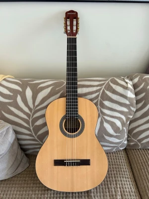 Klassisk gitarr från GECKO - Säljer en klassisk akustisk gitarr från GECKO med naturlig träfärg och mörkbrunt huvud. Gitarren har sex strängar, rundad kropp och dekorativ rosett runt ljudhålet. Perfekt för dig som vill spela allt från klassiskt till pop.