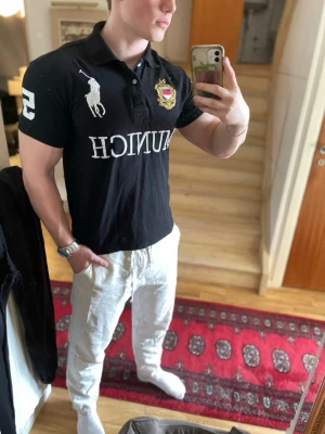 Svart Ralph Lauren Munich piké - Säljer en svart t-shirt från Ralph Lauren med stort MUNICH tryck på bröstet och broderad logga. T-shirten har korta ärmar, krage och knappslå samt en stor vit siffra 5 på ärmen. Perfekt för dig som gillar klassisk och sportig stil.