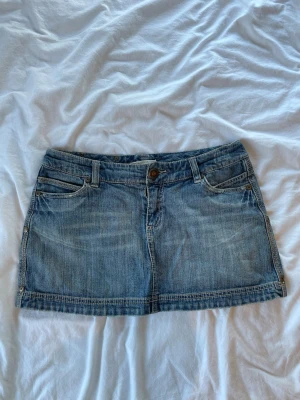 Blå jeanskjol med fickor med detalj - Säljer en blå jeanskjol i kort modell med broderade blommor på bakfickorna. Kjolen har klassiska fem fickor, bälteshällor och knappstängning fram. Perfekt för dig som gillar en avslappnad och trendig stil.