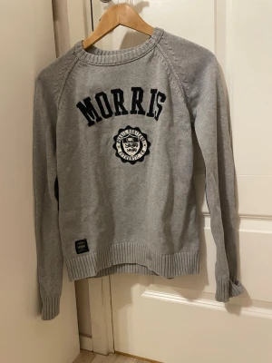 Grå stickad tröja från Morris - Snygg grå stickad tröja från Morris med svart broderad logga och emblem på bröstet. Tröjan har rund halsringning, långa ärmar och ribbade muddar vid ärmslut och nederkant. Perfekt för en stilren och avslappnad look.