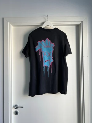 Limited Edition Quavo X BoohooMAN Top - Cool Quavo limited edition 2020 svart t-shirt från Quavo X boohoo MAN collab collectionen med ett stort grafiskt tryck i blått och rosa på ryggen och mindre tryck på bröstet. T-shirten har rund hals och korta ärmar. Perfekt för dig som gillar streetwear och unika prints. 100% bomull. Storlek M / medium. Måtten finns. 