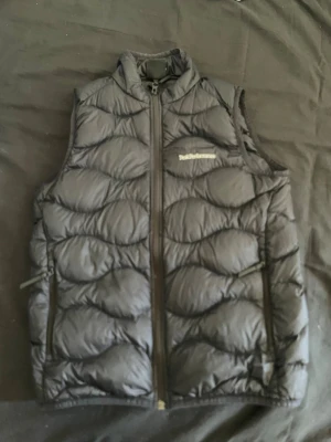 Svart dunväst från Peak Performance - Snygg svart dunväst från Peak Performance med hög krage och dragkedja framtill. Västen har två sidofickor med dragkedja och ett vågigt quiltat mönster. Perfekt för lager-på-lager och håller dig varm under kyliga dagar.