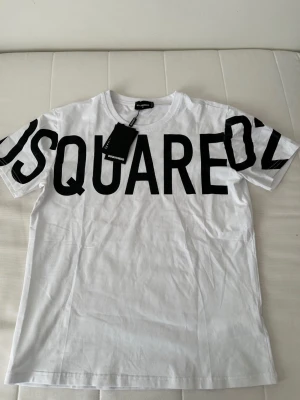 Vit DSQUARED2 t-shirt med tryck - Snygg vit t-shirt från DSQUARED2 med stort svart tryck över bröstet och ärmarna. Klassisk rund halsringning och korta ärmar. Tillverkad i mjuk bomull som känns skön mot huden. Perfekt för en trendig och avslappnad stil.
