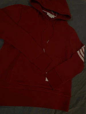 Thom Browne hoodie - Snygg vinröd hoodie från Thom Browne med klassiska vita ränder på ena ärmen och en vit patch med logga på framsidan. Hooiden är i storleken 2 som motsvarar S/M. Hoodien har huva med dragsko och en stor magficka. Perfekt för dig som vill ha en stilren och trendig look. PRISET GÅR ATT DISKUTERA!