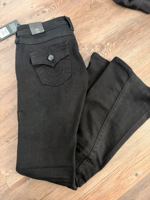 Svarta True Religion jeans - Säljer ett par svarta low rise jeans från True Religion med klassisk ficka och knappdetalj bak. Jeansen har prislappen kvar då dom ej passade mig. Köpta för 1299kr.