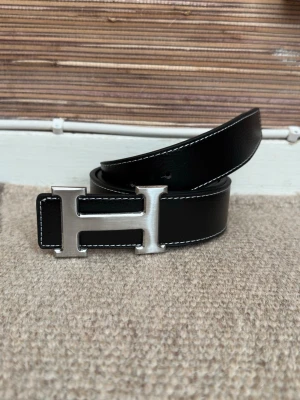 Svart skärp med H-spänne - Stilrent svart skärp med ett ikoniskt H-formad spänne i silver. Skärpet är tillverkat i skinn och har vita kontrastsömmar längs kanterna. Perfekt för att lyfta din outfit med en lyxig touch.