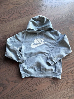 Grå hoodie från Nike med logga - Snygg grå hoodie från Nike med stor vit logga på bröstet. Tröjan har huva, ribbade muddar och en klassisk känguruficka framtill. Perfekt för dig som gillar sportig och avslappnad stil. Materialet är mjukt och bekvämt, idealiskt för chill eller träning.