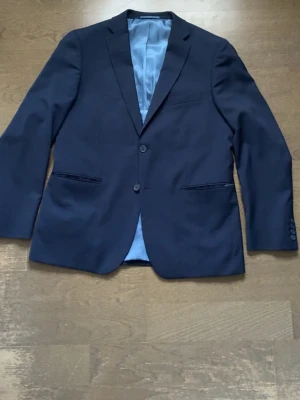 Mörkblå kavaj med två knappar - Snygg mörkblå kavaj med klassisk passform och två knappar framtill. Kavajen har fyra knappar på ärmsluten, två fickor med lock och en bröstficka. Fodret är ljusblått och materialet känns slätt och stilrent.