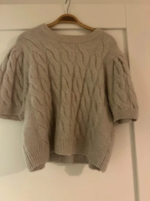 Beige kabelstickad tröja Gina Tricot - Säljer en beige kabelstickad tröja från Gina Tricot med korta puffärmar och rund halsringning. Tröjan har ett snyggt flätmönster och ribbade muddar vid ärmslut och nederkant. Perfekt för dig som gillar en mysig och trendig look.