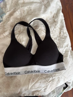 Svart sport-bh från Calvin Klein - Svart sport-bh från Calvin Klein med bred vit resår och logga runt om. Bh:n har korsade axelband i ryggen och är tillverkad i mjukt stretchigt material, perfekt för en aktiv vardag. Enkel och stilren design som ger bra stöd.